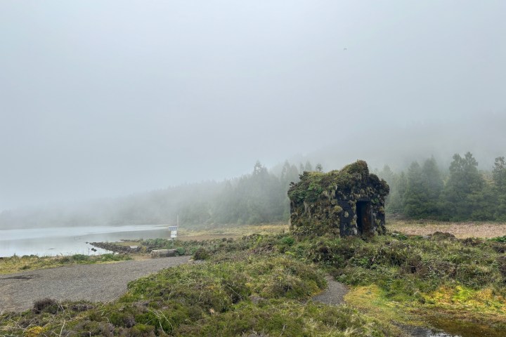 Foggy landscape with a small stone hut, lake, and trees in the background.