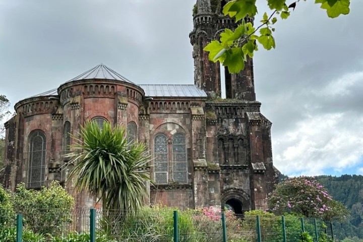 Old stone church with a tall spire, surrounded by greenery and cloudy skies.