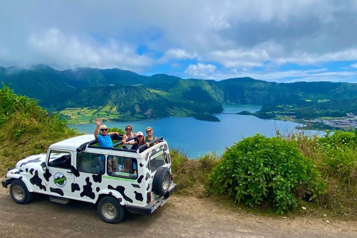 People in cow-print jeep on hill, overlooking lush landscape and lake under cloudy sky.