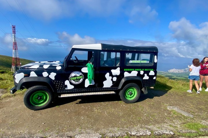 Jeep with cow print parked on a hilltop, two people standing nearby, overlooking a landscape.