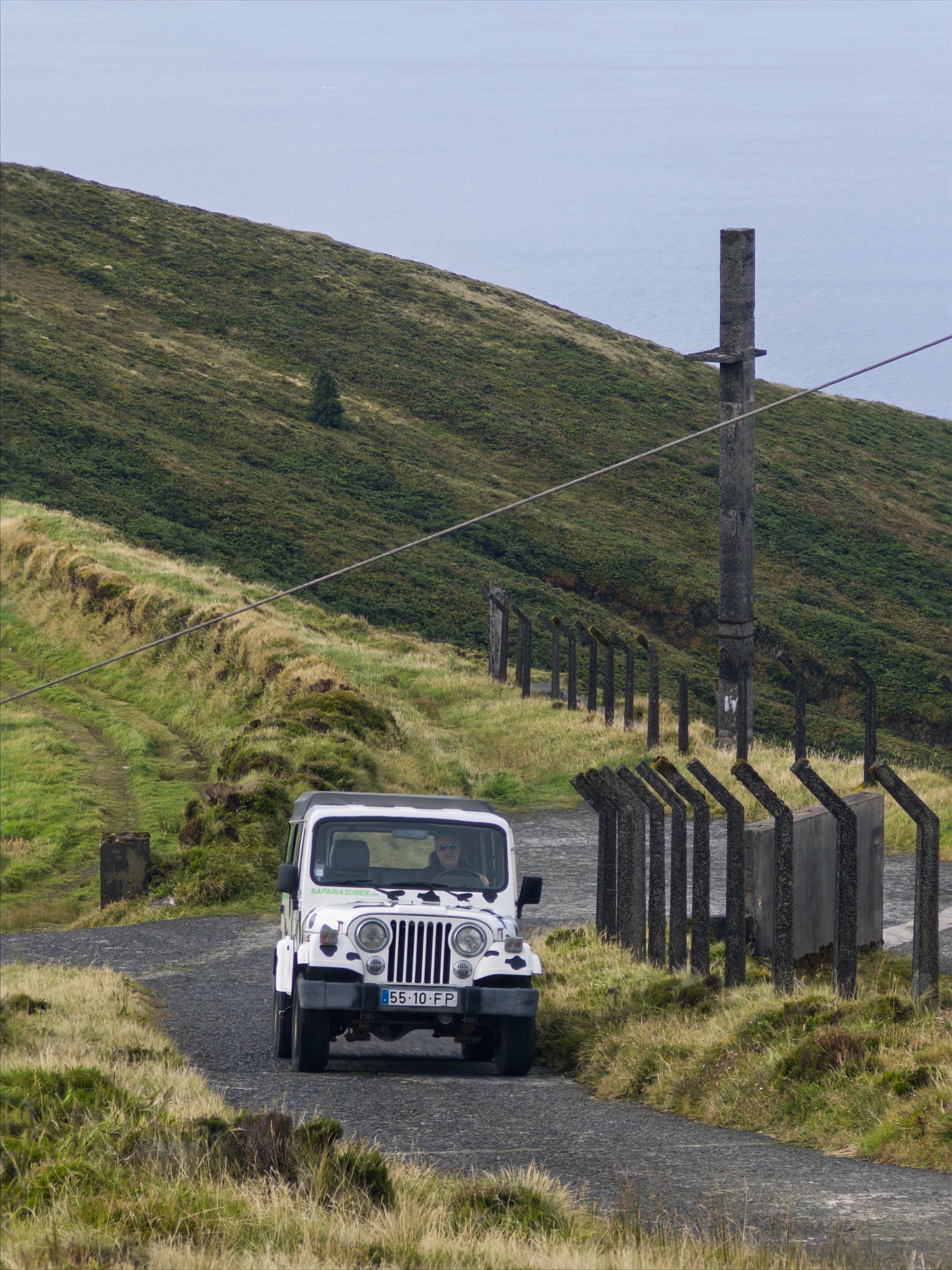 White jeep driving on a rural road with hills and a power line post nearby.