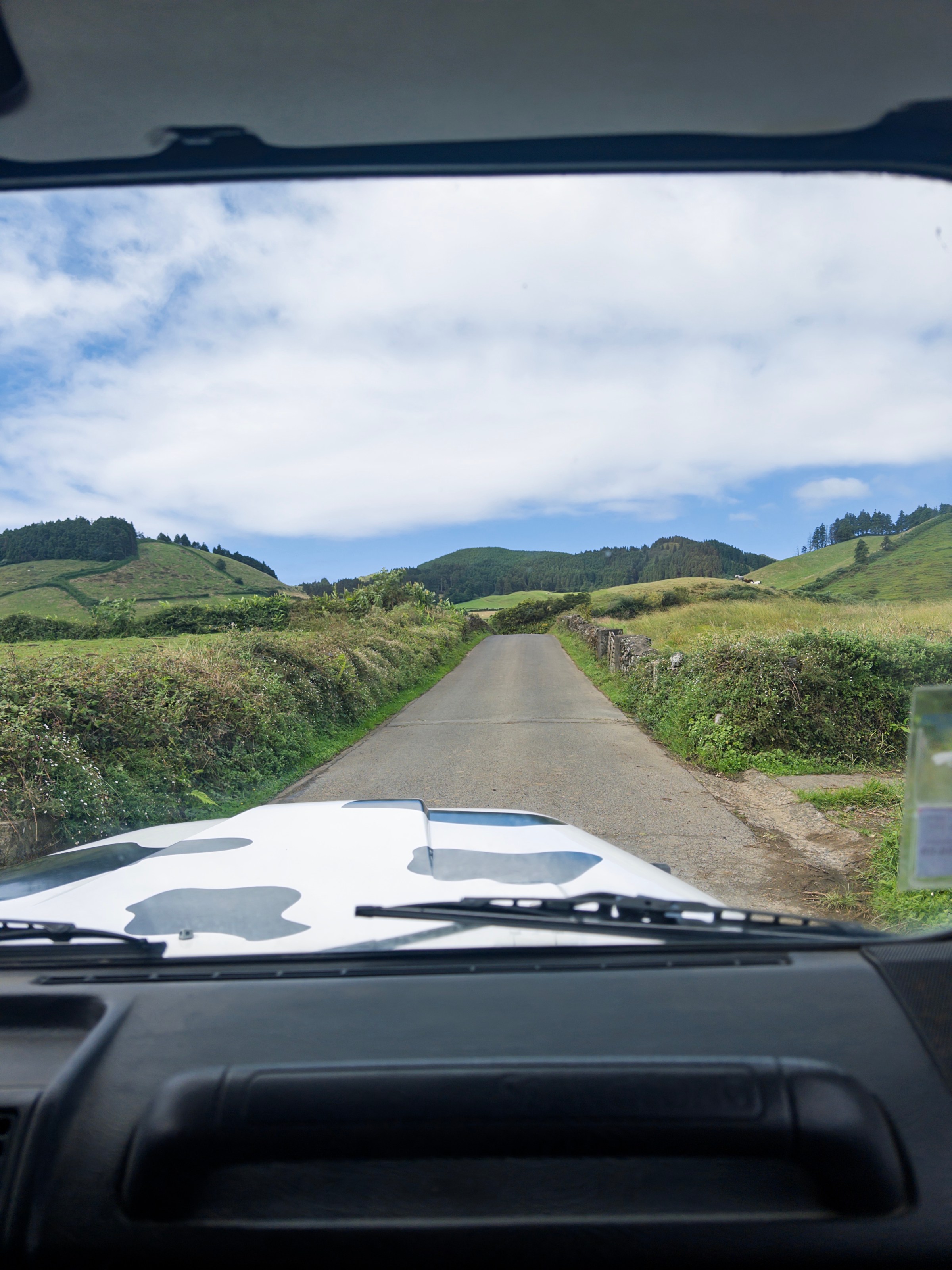 View from car interior of narrow road through green hills under a partly cloudy sky.
