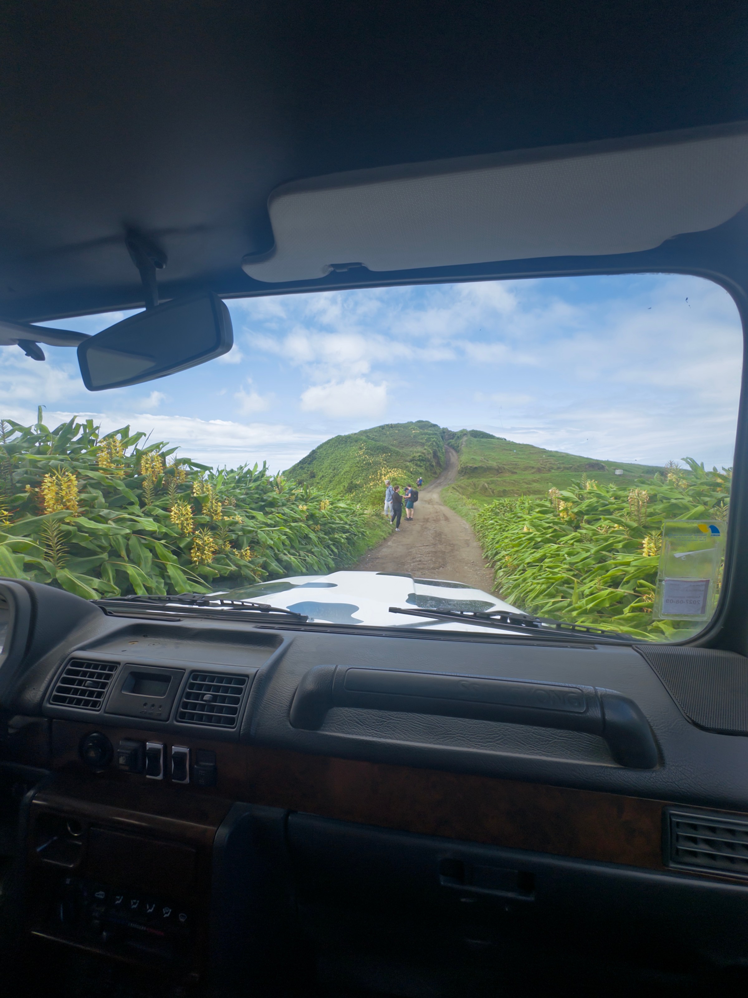View of a dirt road through a car windshield with people walking and greenery outside.