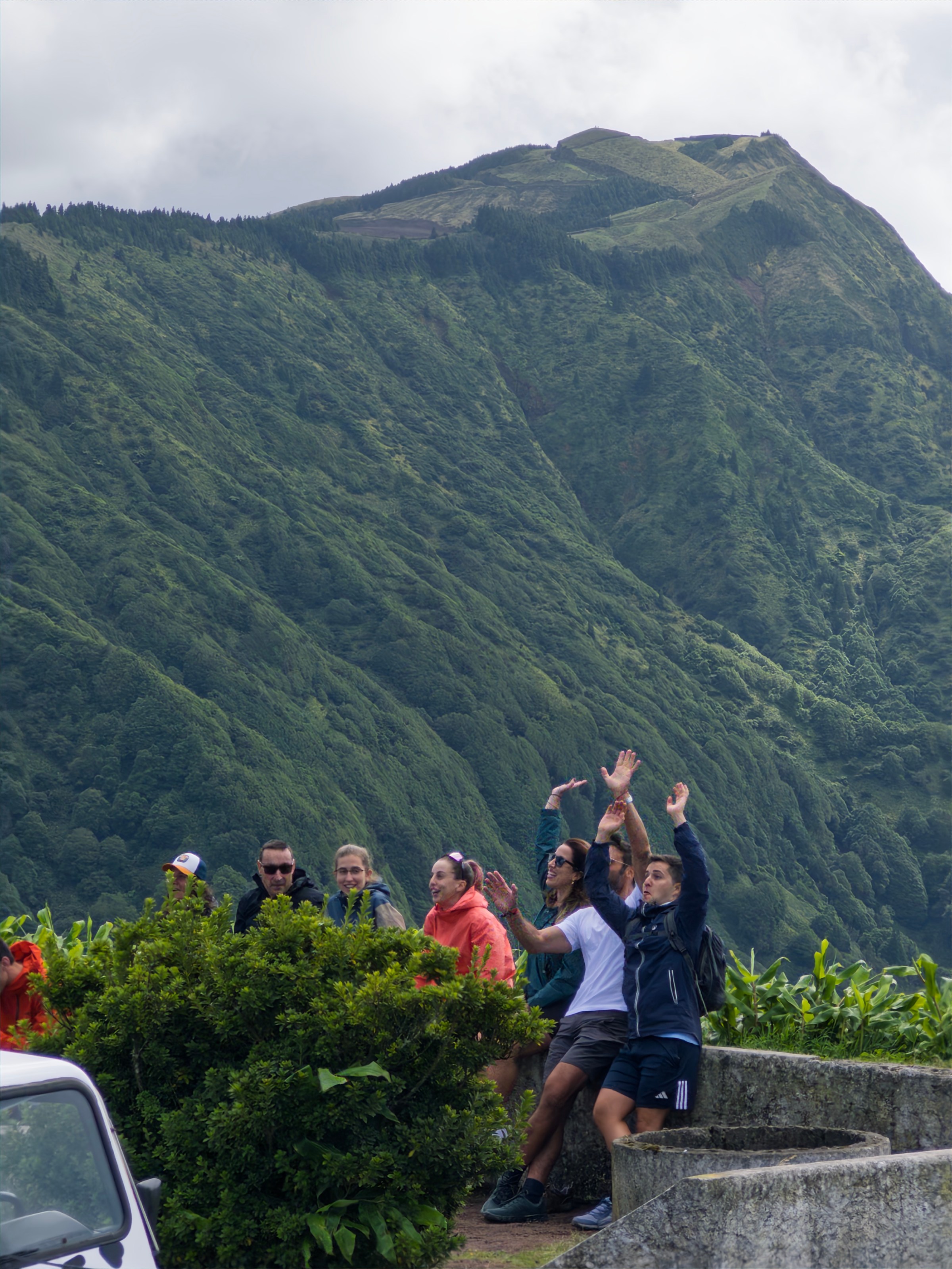 Group posing energetically in front of lush green mountain, cloudy sky above.