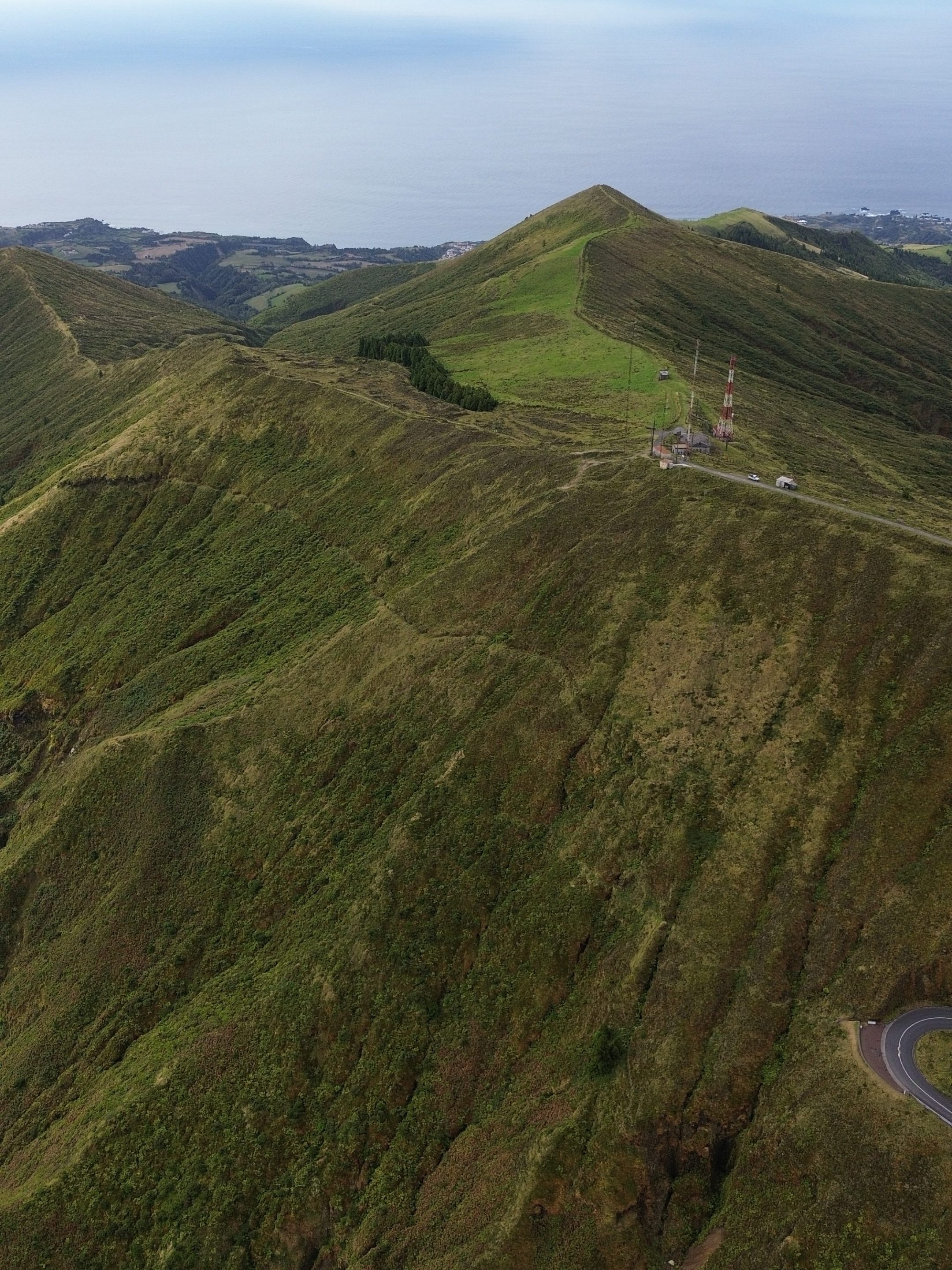 Aerial view of lush green hills with a road, antennas, and ocean in the background under a cloudy sky.