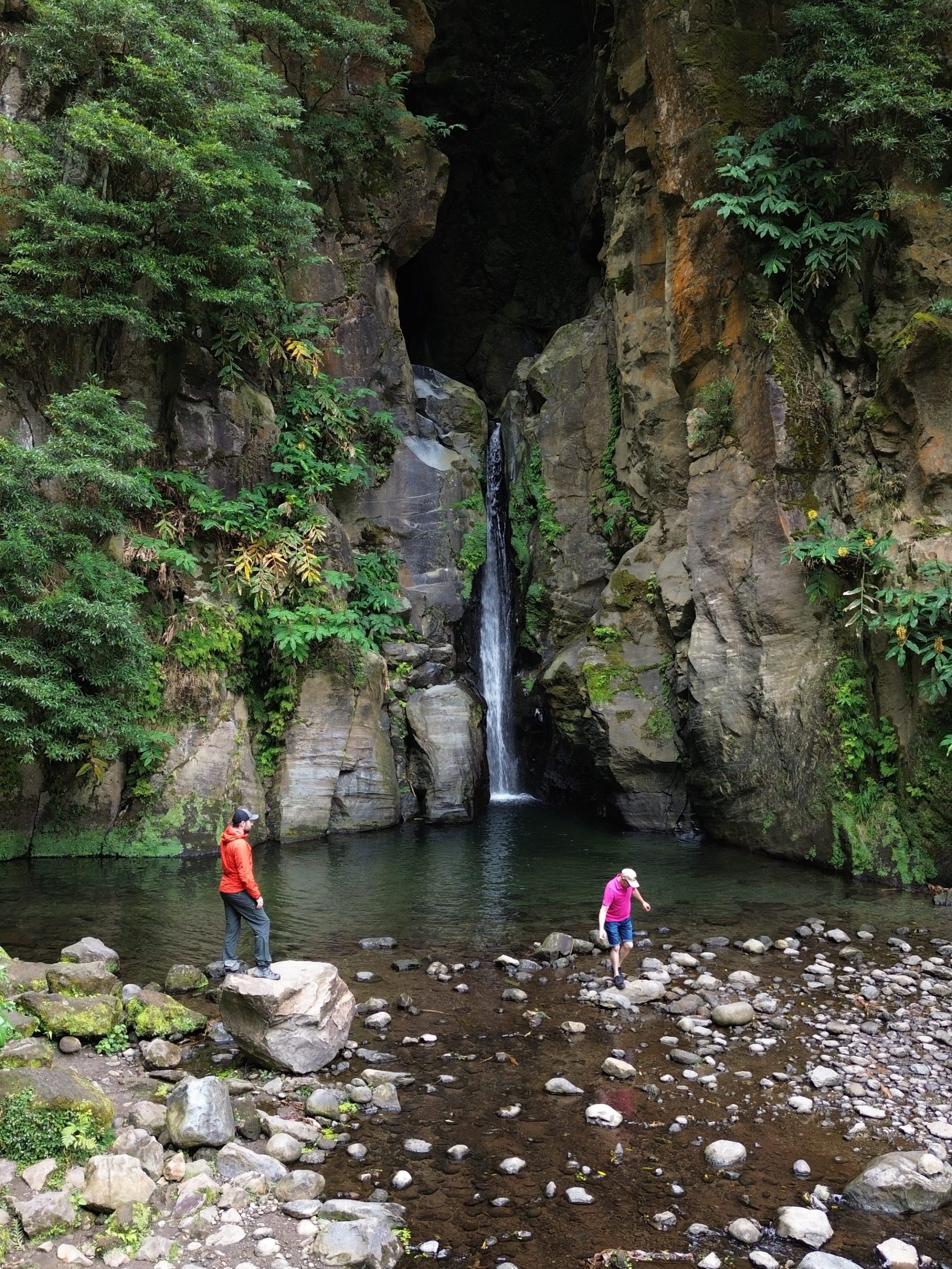 Two people near a small waterfall surrounded by tall, rocky cliffs and greenery.