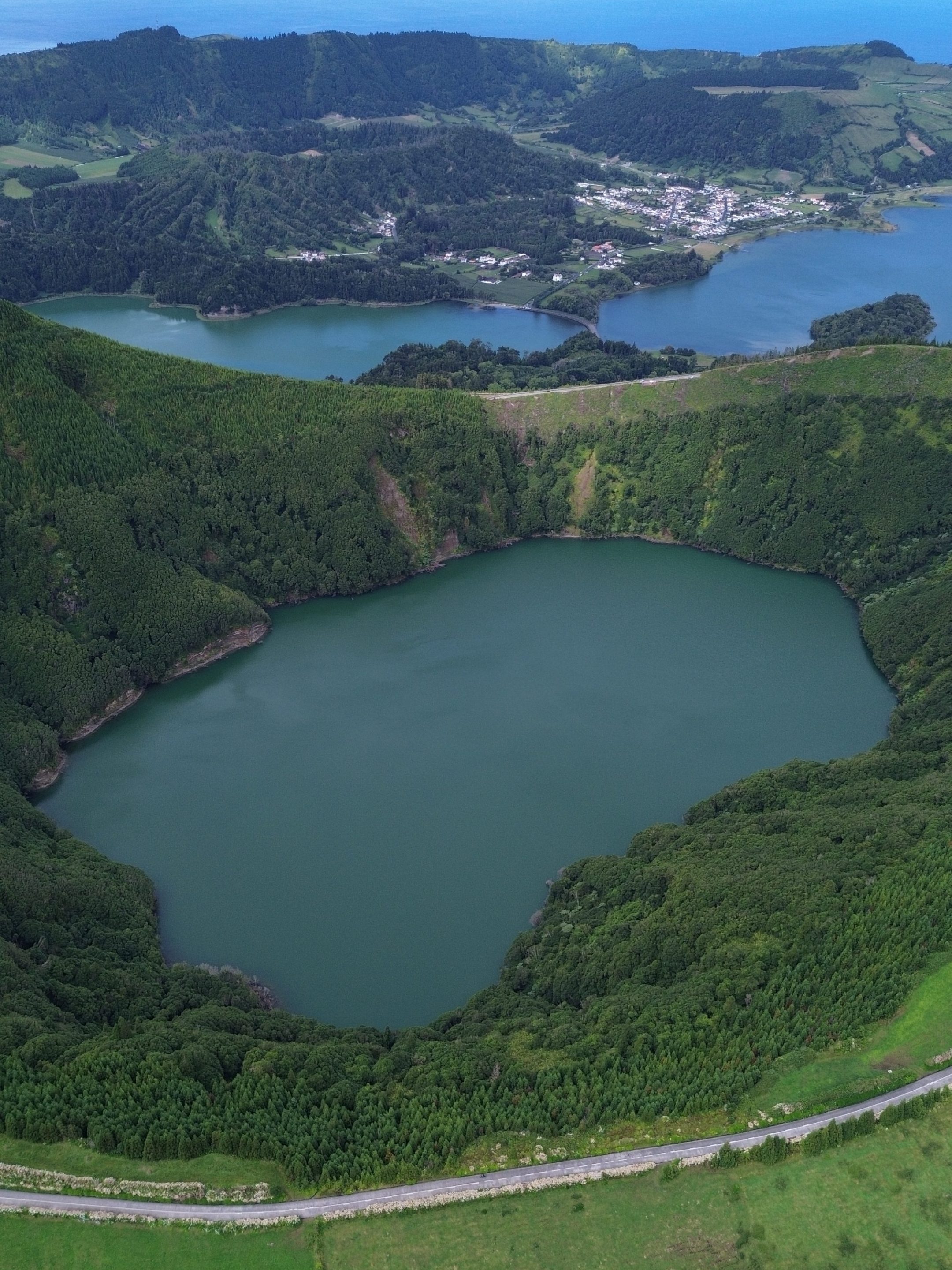 Aerial view of a green crater lake surrounded by lush hills, with another lake and ocean in the background.