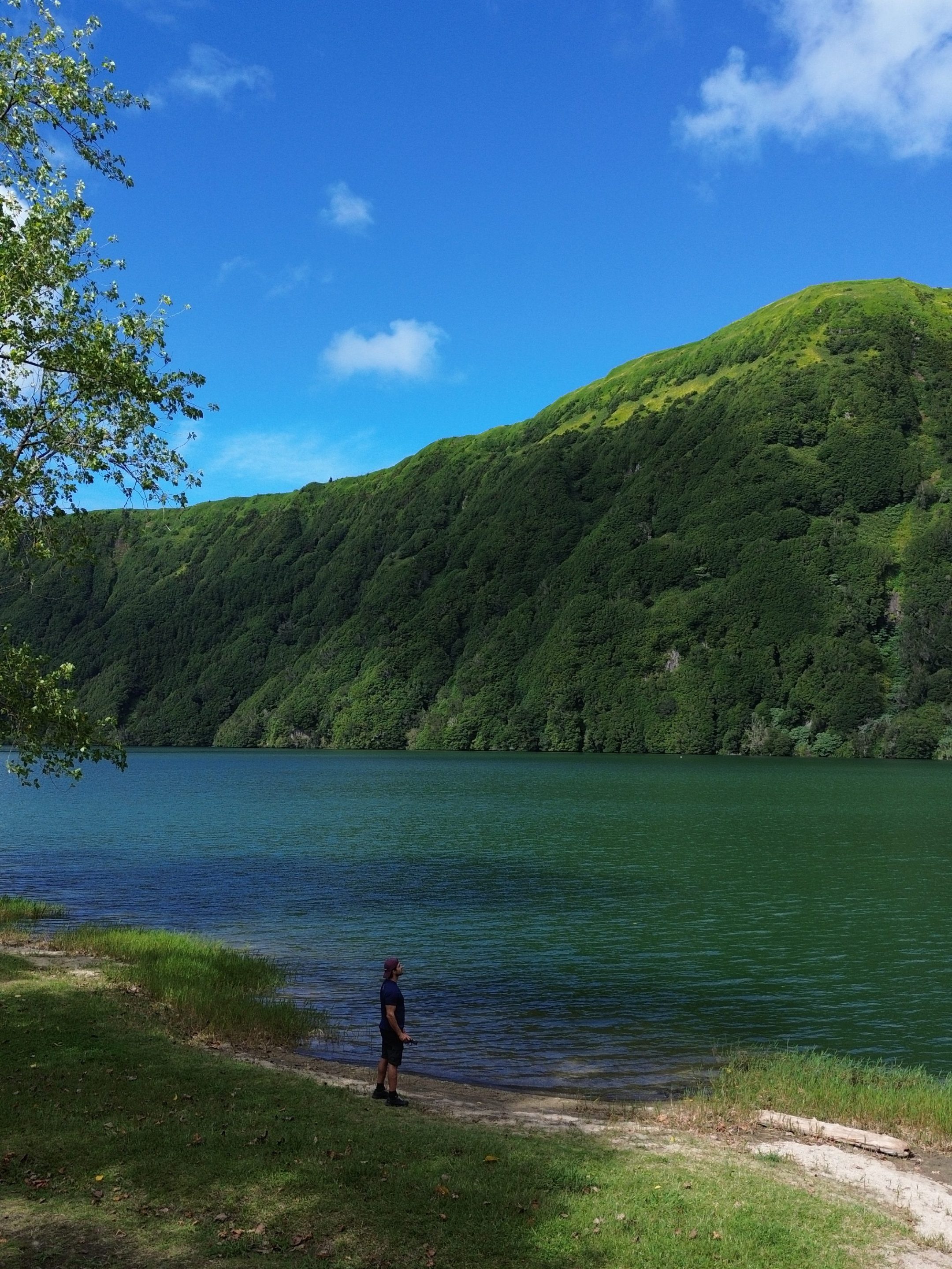 Person standing by a lake with green hills and blue sky in the background.
