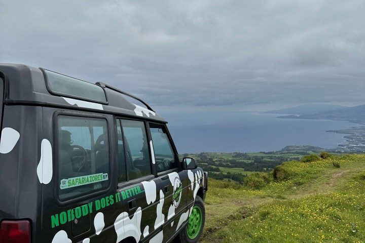 Camouflaged vehicle on grassy hilltop overlooking ocean under cloudy sky.